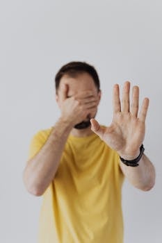 pexels photo 4379960 4379960 Man in yellow shirt covering face with one hand while showing a stop gesture with the other.